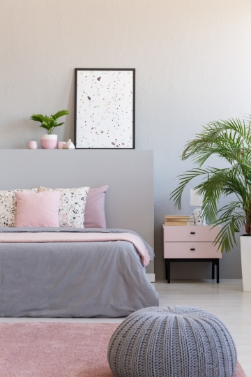 Grey pouf next to bed with cushions in modern bedroom interior with poster and plants. Real photo