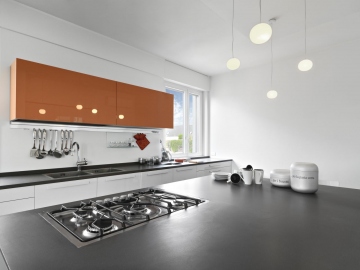 interiors of a modern kitchen to the left the orange wall cabinets in the foreground the gas hob on the background on the worktop of the island kitchen a pile of dishes